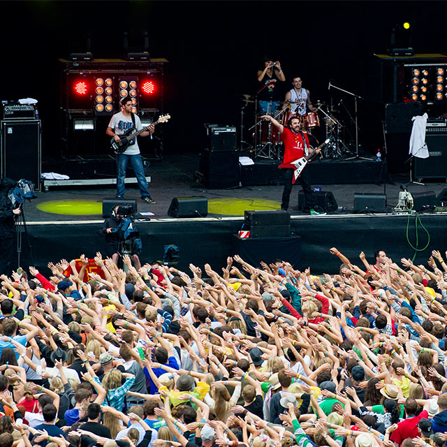Un groupe joue sur une scène en arrière-plan, au premier plan on voit une foule les bras en l’air.