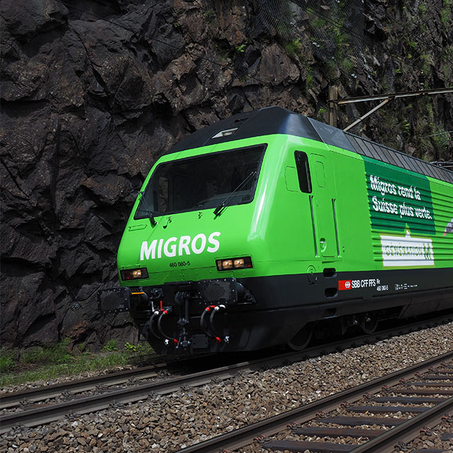 Une locomotive Migros verte qui passe devant un rocher