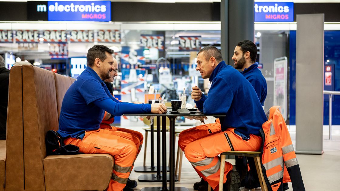 Vier Besucher mit orangen Hosen und blauen Pullovern trinken Kaffee im Migros-Restaurant am Limmatplatz in Zürich